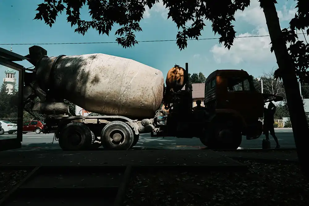 A cement mixer truck parked on the roadside, partially shaded by tree branches.